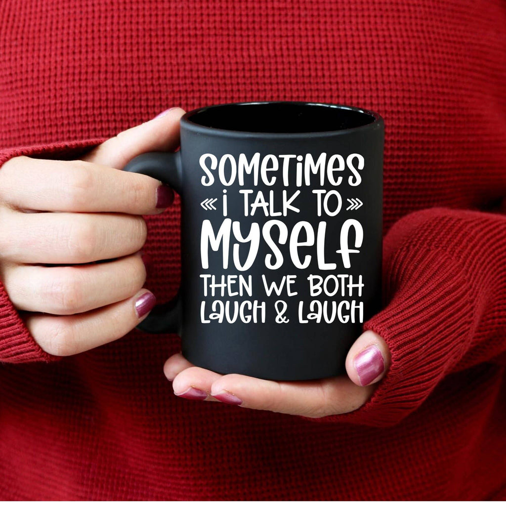 A person in a red knit sweater holds a black ceramic mug with white text that reads, 'SOMETIMES << i TALK TO >> MYSELF THEN WE BOTH LAUGH & LAUGH.'.