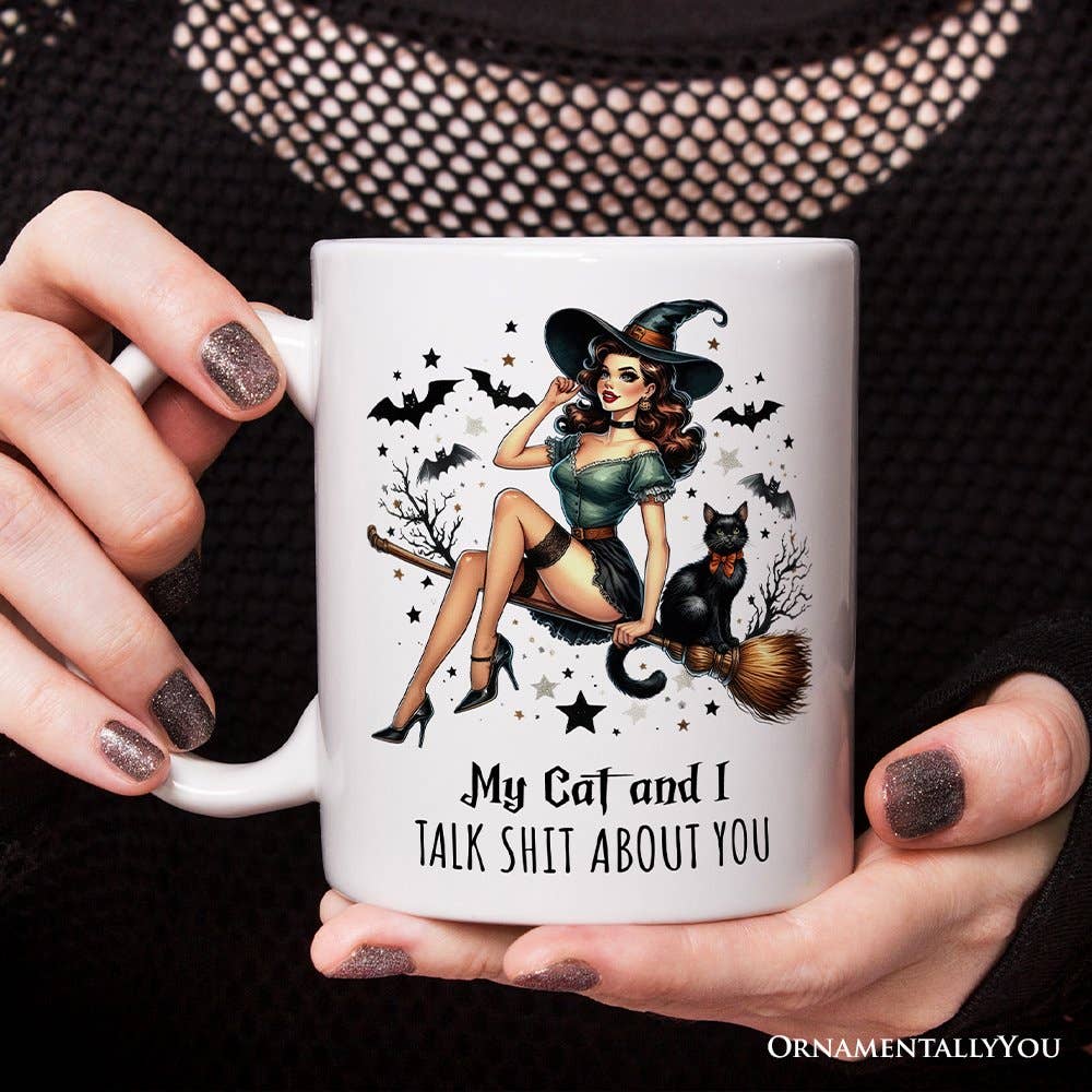 A person holds a white ceramic mug with a vintage-style witch illustration and the text 'My Cat and I TALK SHIT ABOUT YOU'. The person's hands with glittery nail polish are visible holding the mug against a black textured knit background.