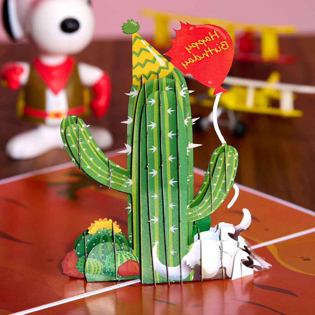 3D paper cactus model with a small white creature on a red surface, with a Snoopy figure in the background.
