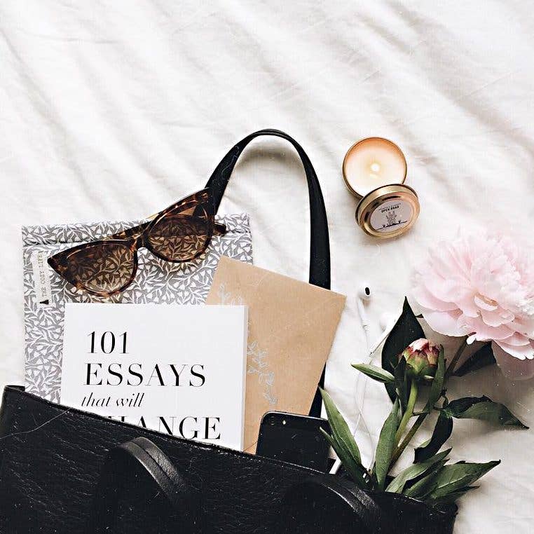 Black handbag with sunglasses, book, phone, and flowers on a white surface