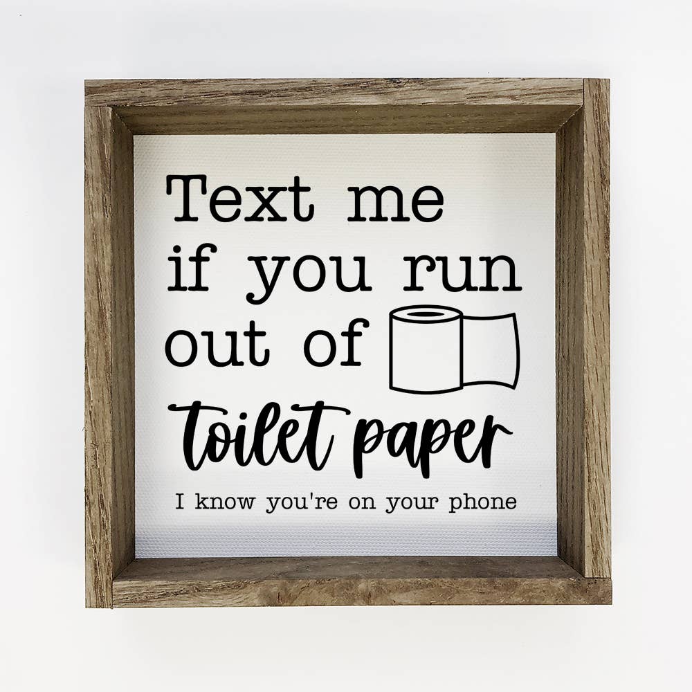 A square framed wall sign with a rustic wood frame and a white background. The sign features black text that reads 'Text me if you run out of toilet paper I know you're on your phone' alongside a simple black line drawing of a toilet paper roll.