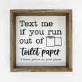 A square framed wall sign with a rustic wood frame and a white background. The sign features black text that reads 'Text me if you run out of toilet paper I know you're on your phone' alongside a simple black line drawing of a toilet paper roll.