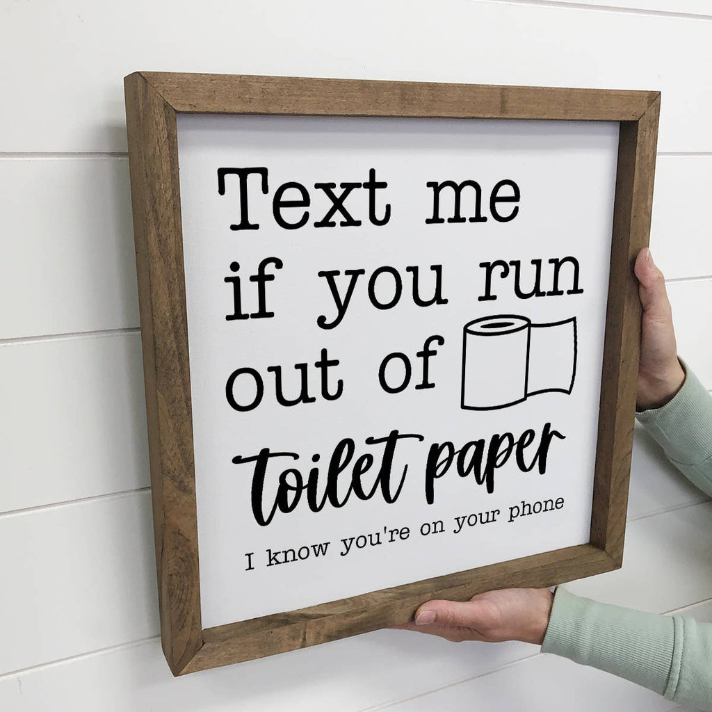 Wooden-framed sign with humorous text about toilet paper, held by a person against a white wall.