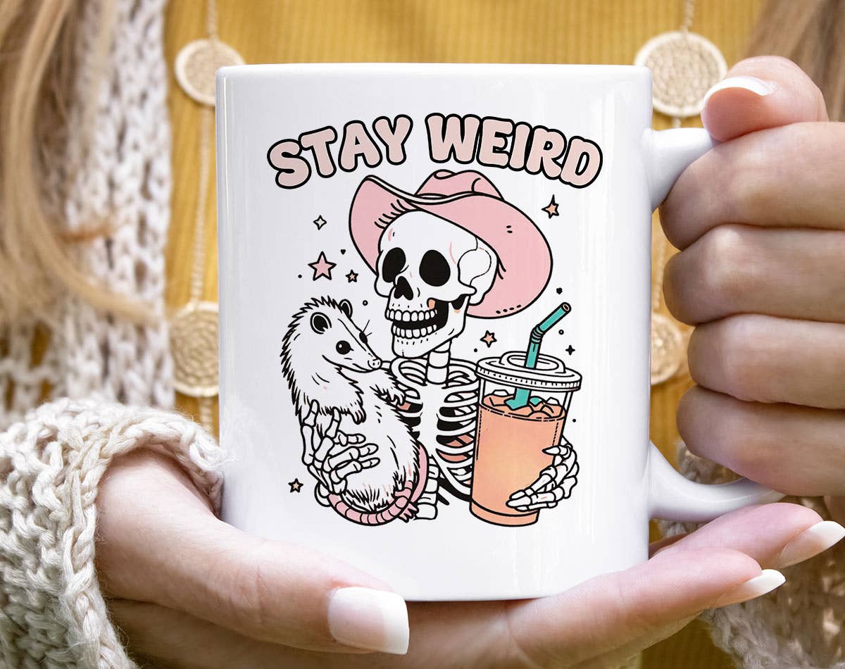 Mug with skeleton and opossum design and 'Stay Weird' text held by a person.