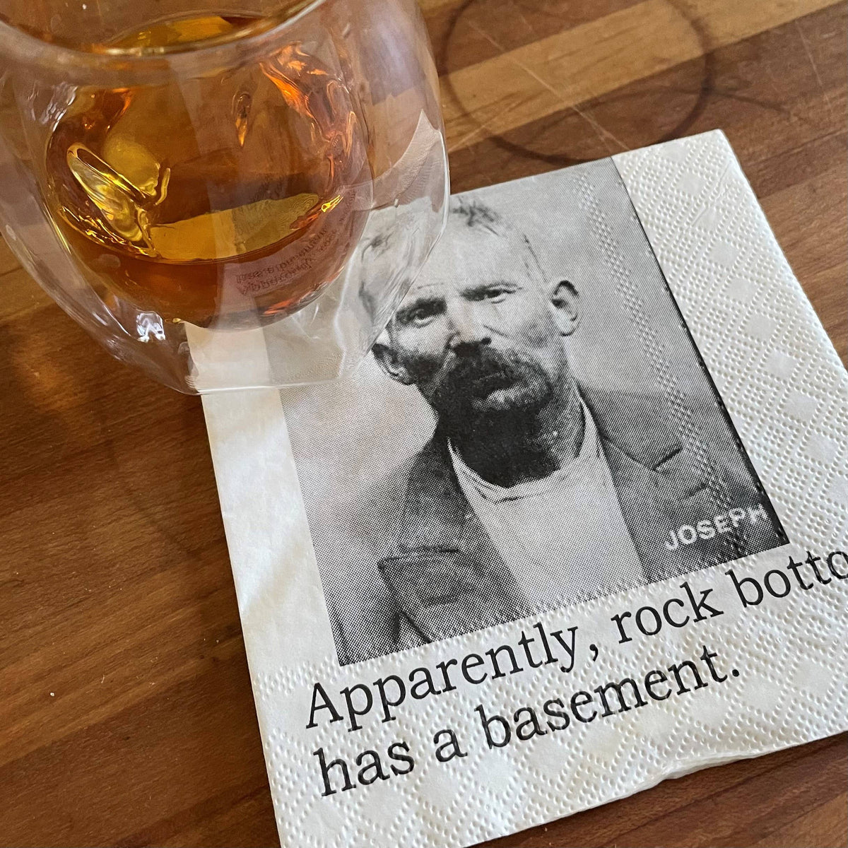 Napkin with a vintage-style portrait and humorous text next to a glass of amber liquid on a wooden surface.