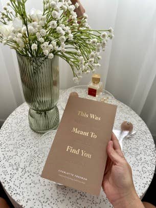 Hand holding a card with text next to a vase of flowers on a table.