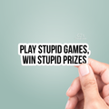 Hand holding a sticker with text 'Play Stupid Games, Win Stupid Prizes' on a light green background.
