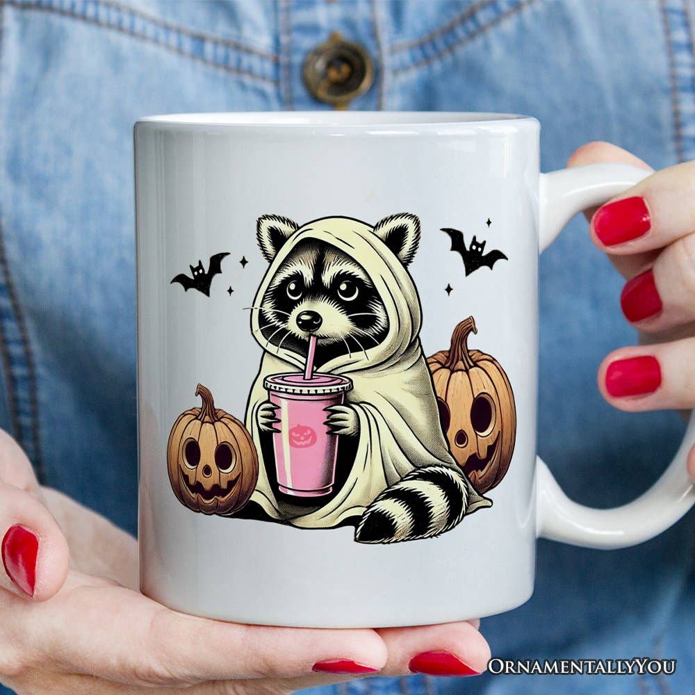 Mug with a raccoon in a hoodie drinking from a pink cup, surrounded by pumpkins and bats, held by a person with red nails.