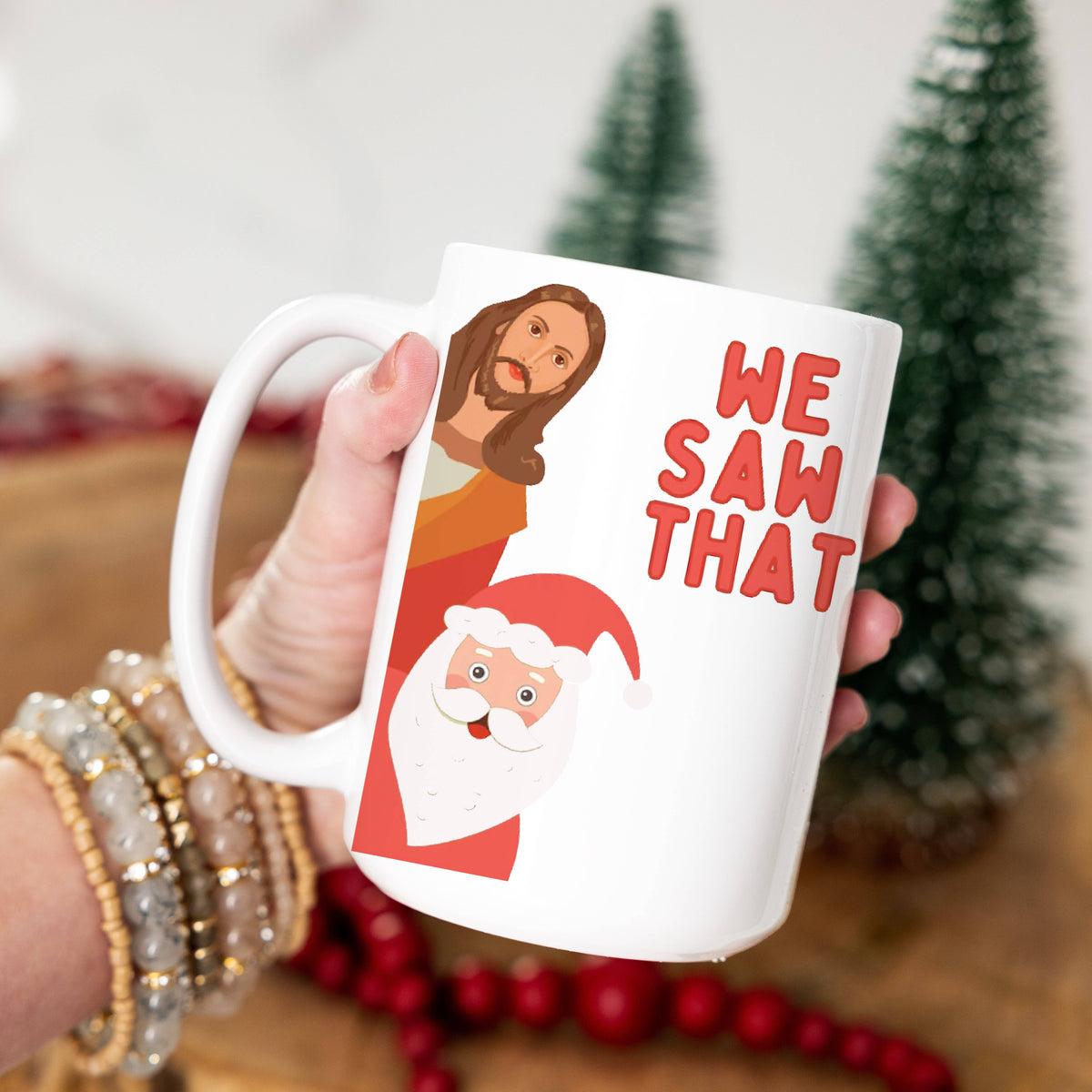 White mug with Christmas-themed design and text held by a person, with a blurred festive background.