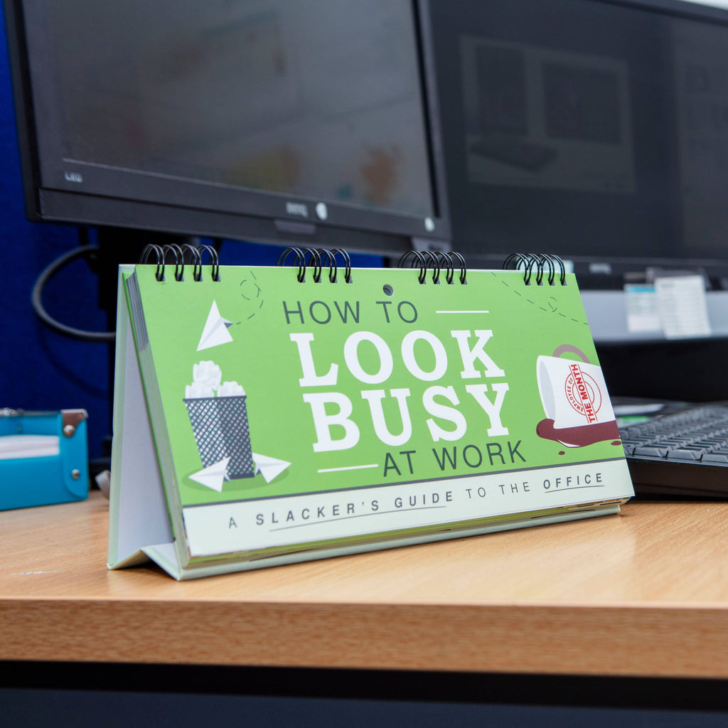 Desk calendar titled 'How to Look Busy at Work' on a desk with computer monitors in the background.