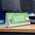 Desk calendar titled 'How to Look Busy at Work' on a desk with computer monitors in the background.