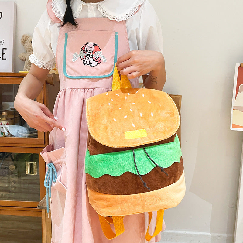 A person wearing a pink dress and apron holds a plush hamburger-shaped backpack with yellow straps, green lettuce layer, brown meat layer, and a tan bun top in a bright indoor room.