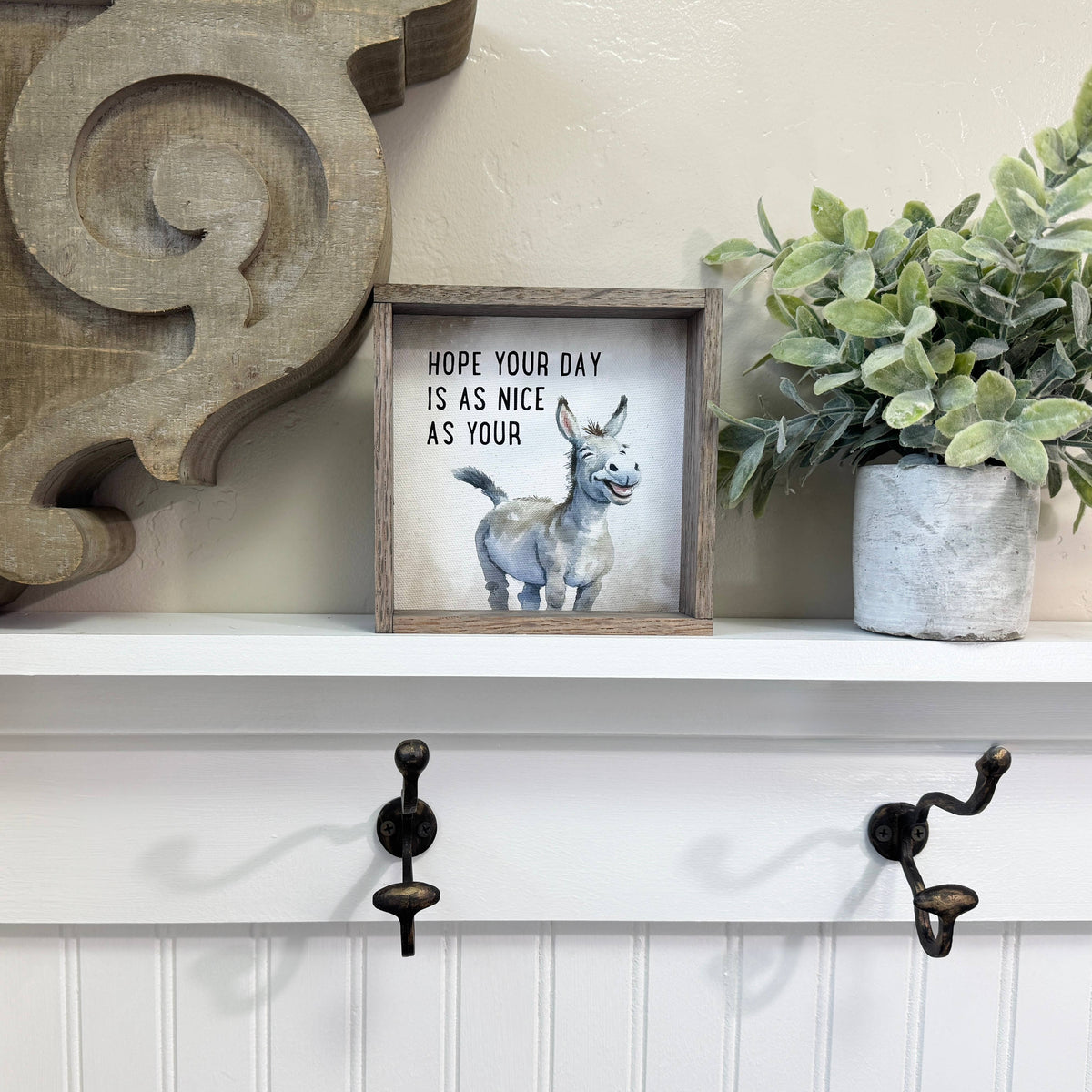 Decorative shelf with a framed picture of a donkey and text, a plant, and wall hooks.