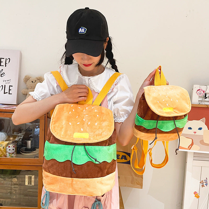 A young girl wearing a black cap and white lace-sleeve top holds two plush hamburger-shaped backpacks with yellow straps in a bright indoor room.
