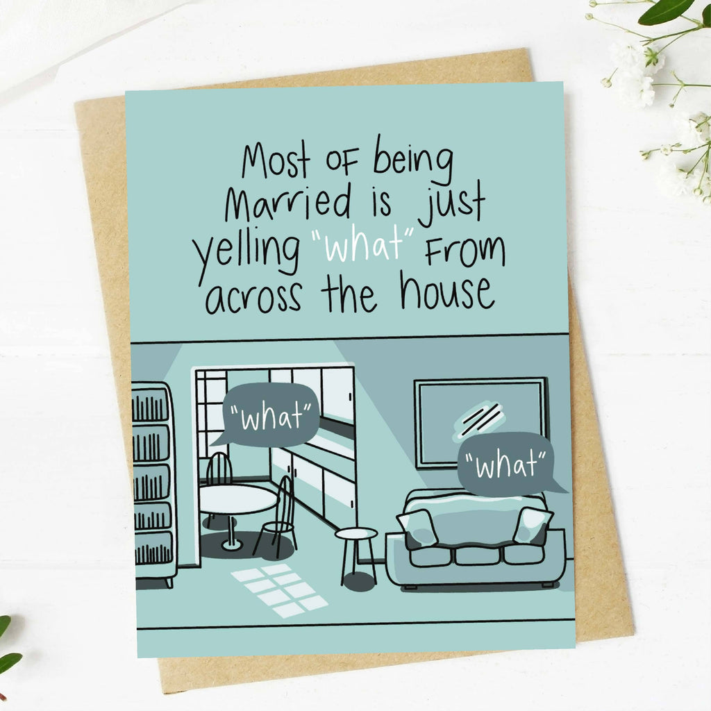 A square greeting card with a light blue background and black handwritten-style text reading, 'Most of being Married is just yelling "what" across the house,' shown with a kraft envelope on a white surface. The lower half of the card includes a simple illustrated living room scene in muted blue-gray tones with speech bubbles also labeled "what.