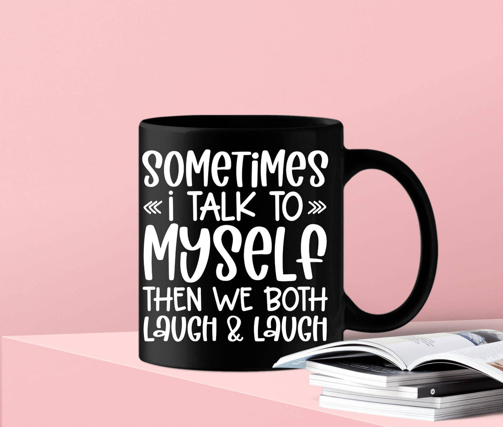 A black ceramic mug with white text reading 'SOMETIMES << i TALK TO >> MYSELF THEN WE BOTH LAUGH & LAUGH' sits on a pink surface next to an open book against a pink background.