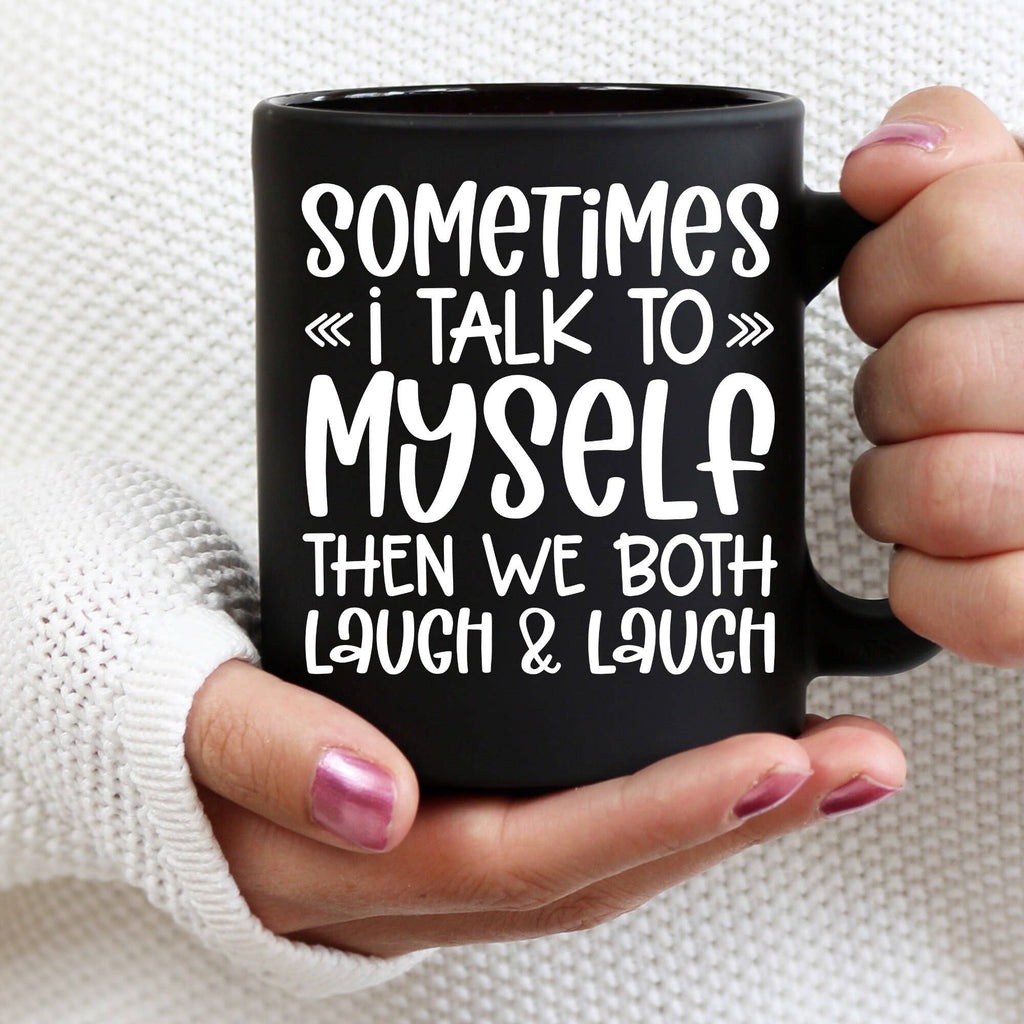 A person holding a black ceramic mug with white text that reads 'SOMETIMES <<i TALK TO>> MYSELF THEN WE BOTH LAUGH & LAUGH'. The mug is held in both hands against a white knit sweater background.
