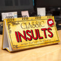 Desk calendar titled 'Classic Insults' on a wooden surface with a blurred office background.