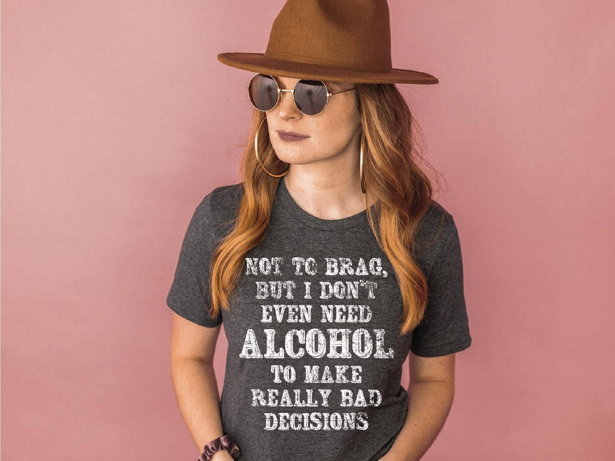 Person wearing a dark gray t-shirt with humorous text, sunglasses, and a brown hat against a pink background