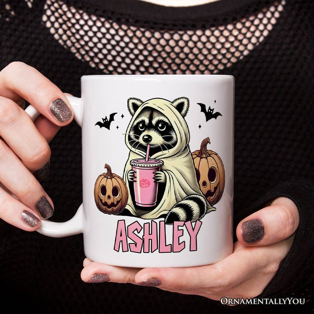 Person holding a white mug with a cartoon raccoon in a hoodie design, surrounded by pumpkins and bats, with 'ASHLEY' printed on it.
