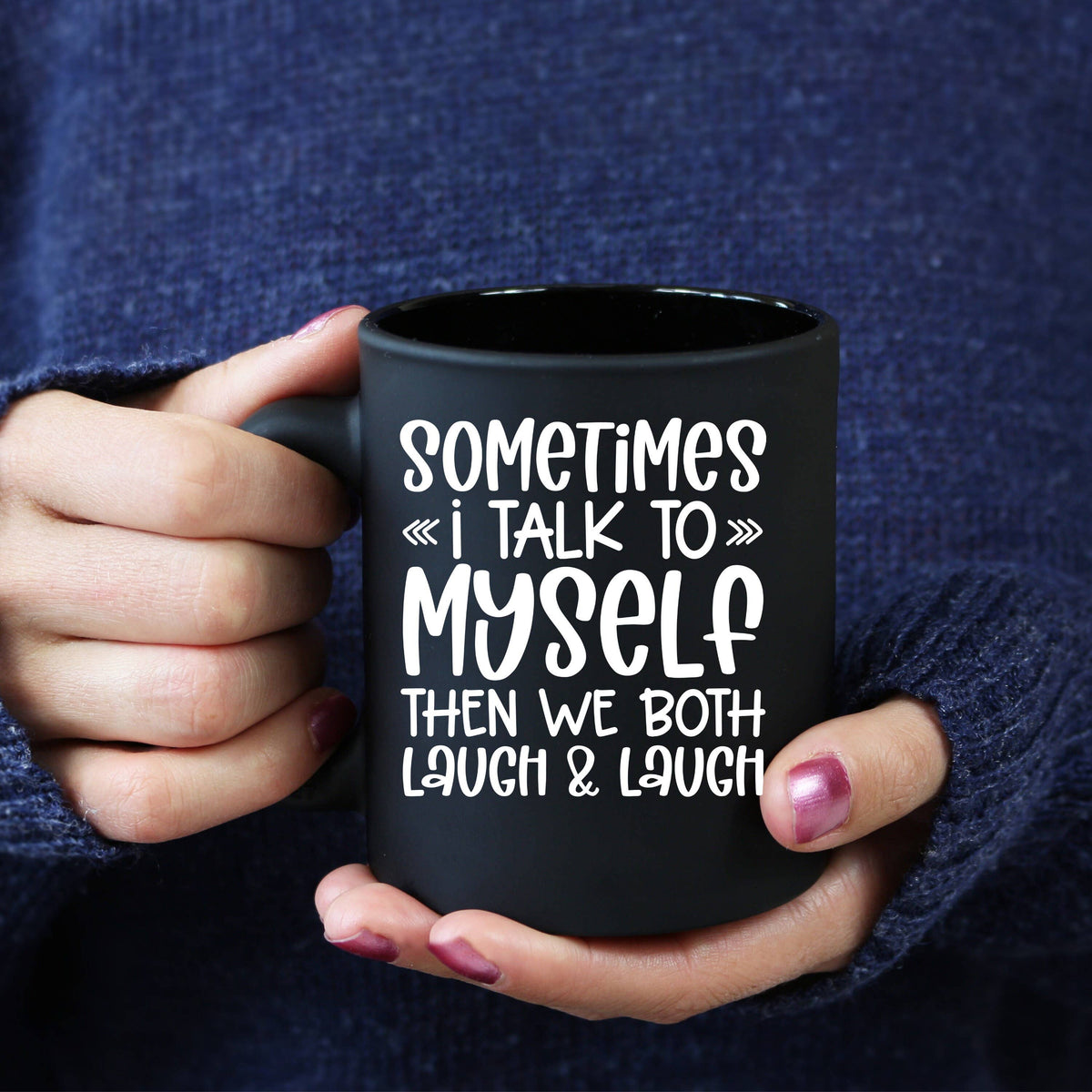 A person holding a black ceramic mug with white printed text that reads 'SOMETIMES I TALK TO MYSELF THEN WE BOTH LAUGH & LAUGH'. The person is wearing a dark blue knit sweater and has pink manicured nails.